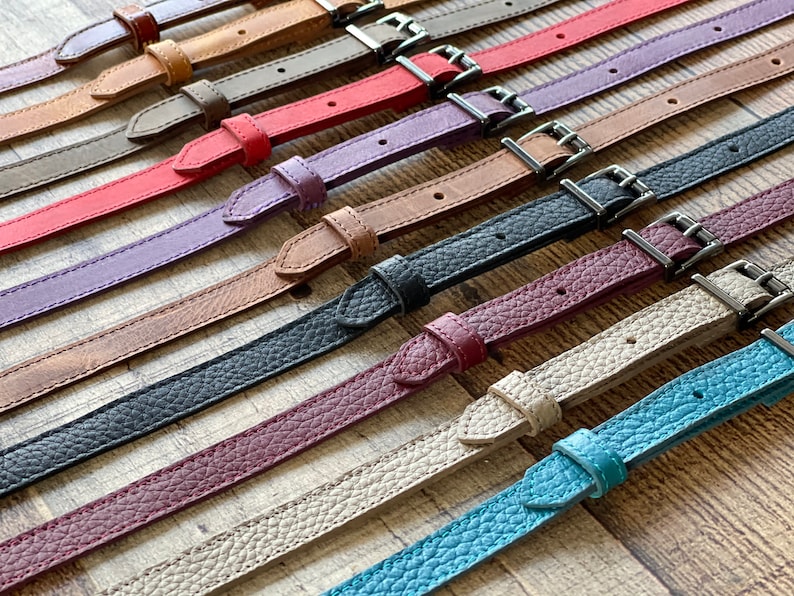 May include: A collection of thin leather straps in various colors including brown, tan, gray, red, purple, black, burgundy, beige, and teal. Each strap features a matching leather keeper, a pointed tip, and a silver-toned metal buckle.