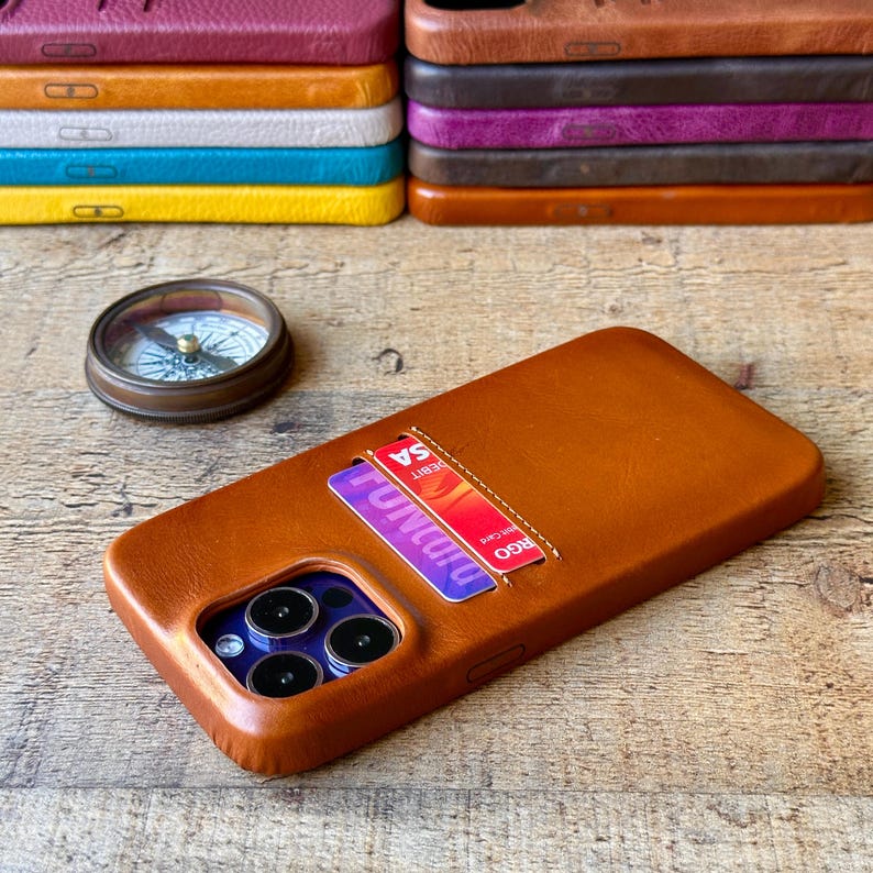 Peut inclure: Une coque de t&eacute;l&eacute;phone en cuir beige avec un porte-cartes contenant deux cartes bancaires, dot&eacute;e d'une d&eacute;coupe pour trois objectifs photo. Une boussole ronde en laiton est pos&eacute;e &agrave; c&ocirc;t&eacute;. En arri&egrave;re-plan, une pile de coques en cuir est visible.