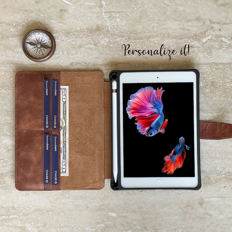 May include: A brown leather iPad case with a black frame, a white stylus, and a screen displaying two colorful betta fish. The case has card slots and a $50 bill. A compass is on the left, and the text "Personalize it!" is visible.