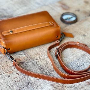 May include: A rectangular tan leather bag featuring a top handle with a metal rivet, a detachable shoulder strap with metal clasps, and a dark zipper closure. A small circular compass is positioned near the bag on the surface.
