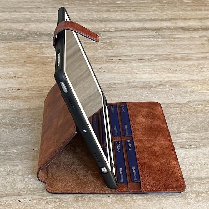 May include: A brown leather tablet case with a black frame, featuring a magnetic closure strap. The case is designed to hold a tablet upright, with slots for cards visible inside. The leather has a textured appearance.