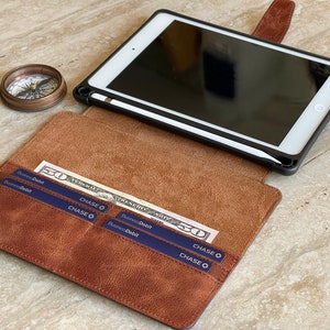 May include: A brown leather iPad case with a black frame, holding a white iPad and a stylus. The case is open, revealing a pocket with a fifty-dollar bill and Chase debit cards. A vintage compass sits nearby.