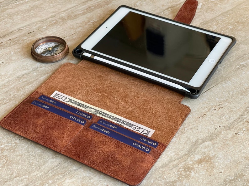 May include: A brown leather iPad case with a black frame, holding a white iPad and a stylus. The case is open, revealing a pocket with a fifty-dollar bill and Chase debit cards. A vintage compass sits nearby.