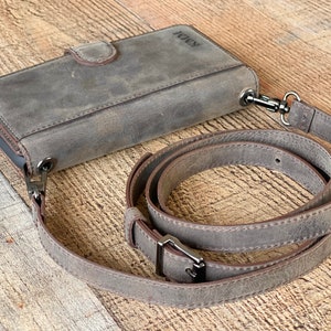 May include: A grey leather phone wallet case with a magnetic closure and a detachable, adjustable shoulder strap. The case features a smooth, matte finish and the embossed letters KMM on the top corner. Dark metal hardware connects the strap to the case.