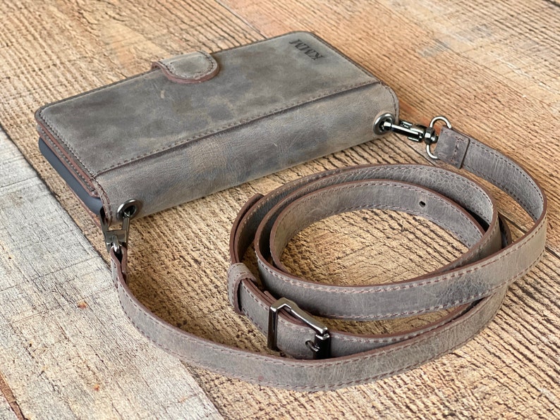 May include: A grey leather phone wallet case with a magnetic closure and a detachable, adjustable shoulder strap. The case features a smooth, matte finish and the embossed letters KMM on the top corner. Dark metal hardware connects the strap to the case.