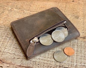 May include: A brown leather wallet with a zipped coin pocket, partially open to show several coins. The wallet is rectangular and has a textured surface. The zip pull is a small leather tab. Several coins are visible, including quarters and a penny.