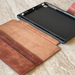 May include: A brown leather tablet case with a black interior frame. The case is open, revealing a light brown interior and card slots. The case has a magnetic closure and is designed to protect the tablet.