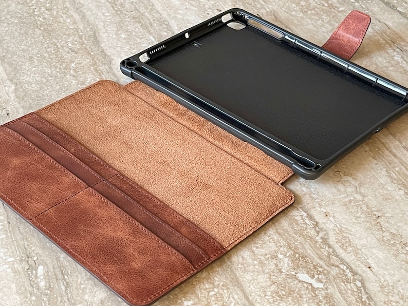 May include: A brown leather tablet case with a black interior frame. The case is open, revealing a light brown interior and card slots. The case has a magnetic closure and is designed to protect the tablet.