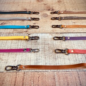Op de afbeelding: Een verzameling leren polsbandjes in diverse kleuren, waaronder turquoise, geel en paars. Elke band heeft een metalen sluiting om aan een tas of ander item te bevestigen. De bandjes liggen op een houten ondergrond.