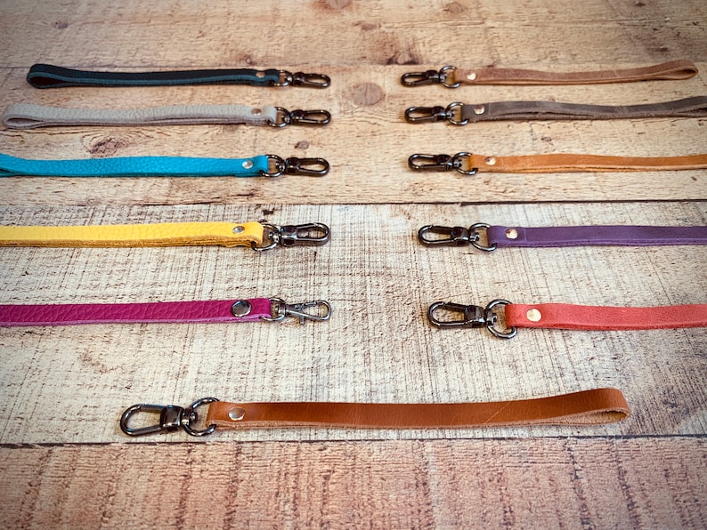 Op de afbeelding: Een verzameling leren polsbandjes in diverse kleuren, waaronder turquoise, geel en paars. Elke band heeft een metalen sluiting om aan een tas of ander item te bevestigen. De bandjes liggen op een houten ondergrond.