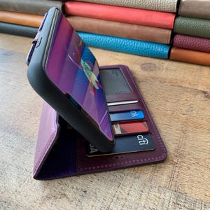 Puede incluir: Una funda de tel&eacute;fono de cuero morado con una cartera incorporada. El tel&eacute;fono muestra una imagen floral colorida. Se ven varias tarjetas de cr&eacute;dito en la cartera. Una pila de art&iacute;culos de cuero de varios colores est&aacute; en el fondo.