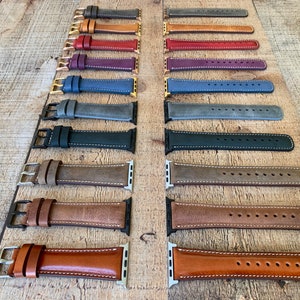 May include: A collection of leather watch bands in various colors, including gray, brown, and blue. Each band features contrasting white stitching and metal hardware. The bands are displayed on a wooden surface.