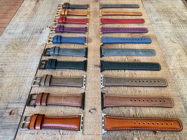 May include: A collection of leather watch bands in various colors, including gray, brown, and blue. Each band features contrasting white stitching and metal hardware. The bands are displayed on a wooden surface.