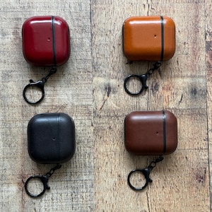 May include: Four square-shaped protective cases for wireless earbuds, each featuring a smooth leather-like finish in red, tan, black, and dark brown. Every case includes a black metal hinge and a matching black circular keychain ring attachment.