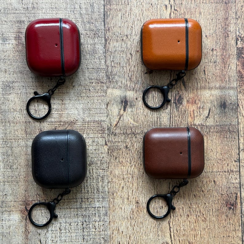 May include: Four square-shaped protective cases for wireless earbuds, each featuring a smooth leather-like finish in red, tan, black, and dark brown. Every case includes a black metal hinge and a matching black circular keychain ring attachment.