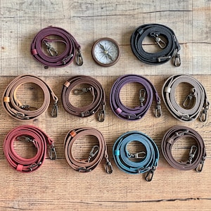 May include: A collection of leather bag straps in various colors, including burgundy, black, tan, purple, and teal, with silver-tone swivel clasps. A vintage-style compass is also present. The straps are arranged on a wooden surface.