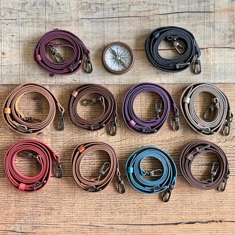 May include: Ten coiled leather bag straps in various colors including burgundy, black, tan, brown, purple, gray, red, light brown, blue, and dark brown, each featuring metal swivel lobster clasps and a small round compass in the center.