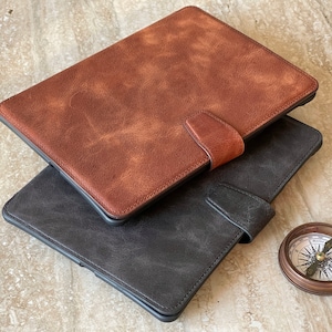 May include: Two leather tablet cases, one brown and one dark gray, are stacked. Each case has a magnetic closure. A small, antique-style compass sits nearby. The cases have a textured, distressed appearance.