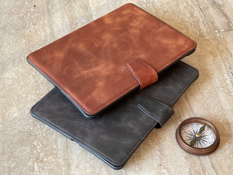 May include: Two leather tablet cases, one brown and one dark gray, are stacked. Each case has a magnetic closure. A small, antique-style compass sits nearby. The cases have a textured, distressed appearance.