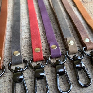 Op de afbeelding: Een verzameling leren polsbandjes in diverse kleuren, waaronder rood, paars en grijs. Elk bandje is bevestigd aan een zwarte metalen sluiting met een goudkleurige klinknagel. De bandjes zijn gerangschikt op een houten oppervlak.