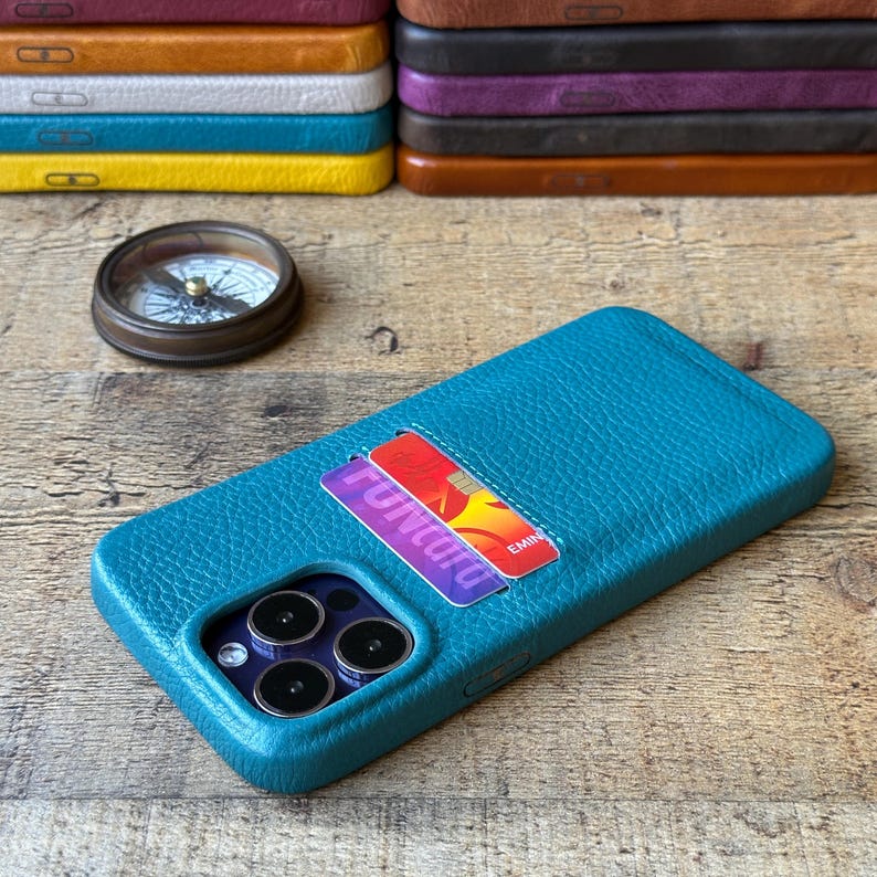 Peut inclure: Une coque de t&eacute;l&eacute;phone en cuir textur&eacute; bleu sarcelle avec un porte-carte contenant une carte bancaire. La coque pr&eacute;sente une d&eacute;coupe pour triple appareil photo. En arri&egrave;re-plan, une pile de coques en cuir color&eacute;es et une boussole ronde.