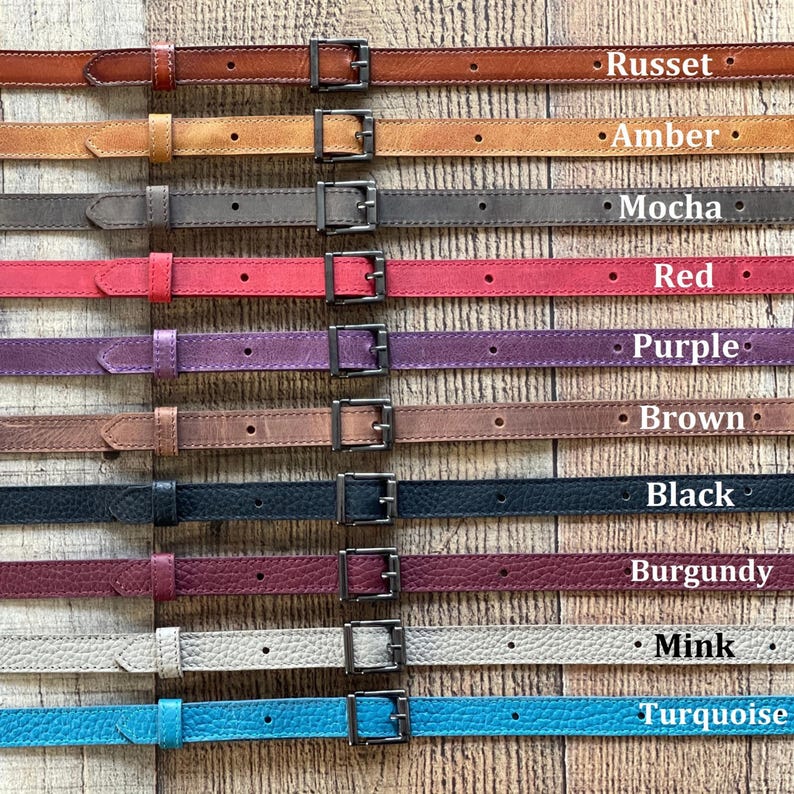 May include: A collection of ten thin leather belts arranged horizontally, each featuring a metal buckle and a single loop. The belts are labeled with the colors Russet, Amber, Mocha, Red, Purple, Brown, Black, Burgundy, Mink, and Turquoise.
