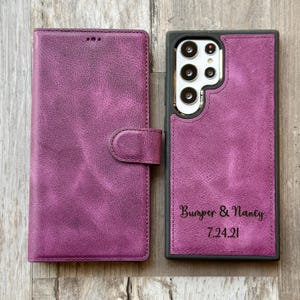 K&ouml;nnte beinhalten: Eine Handyh&uuml;lle aus lila Leder mit schwarzem Rahmen und einer Klappe, die mit einem Knopf geschlossen wird. Die H&uuml;lle tr&auml;gt den Text "Bumper & Nancy 7.24.21" auf der R&uuml;ckseite.