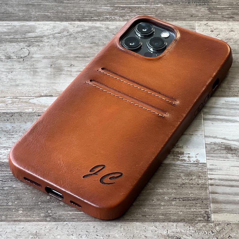 Peut inclure: Une coque de t&eacute;l&eacute;phone en cuir marron clair dot&eacute;e d'une fente pour carte &agrave; l'arri&egrave;re et d'une d&eacute;coupe pour un syst&egrave;me de cam&eacute;ra &agrave; trois objectifs. L'&eacute;tui est grav&eacute; des initiales JC en &eacute;criture cursive pr&egrave;s du bord inf&eacute;rieur.