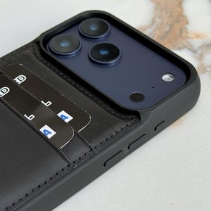 Peut inclure: Une coque de smartphone noire avec un porte-cartes en cuir int&eacute;gr&eacute; au dos. La coque pr&eacute;sente un cadre sur&eacute;lev&eacute; autour du triple capteur photo et contient deux cartes bancaires noires ins&eacute;r&eacute;es dans les fentes, affichant du texte blanc partiel.