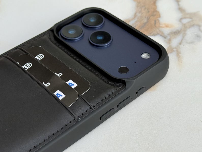Peut inclure: Une coque de smartphone noire avec un porte-cartes en cuir int&eacute;gr&eacute; au dos. La coque pr&eacute;sente un cadre sur&eacute;lev&eacute; autour du triple capteur photo et contient deux cartes bancaires noires ins&eacute;r&eacute;es dans les fentes, affichant du texte blanc partiel.