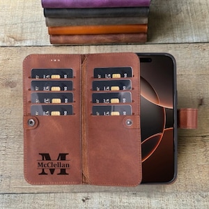 May include: Open brown leather phone case with card slots and a snap closure. The case holds a smartphone and has the name "McClellan" engraved with the letter "M". A stack of leather items is visible in the background.