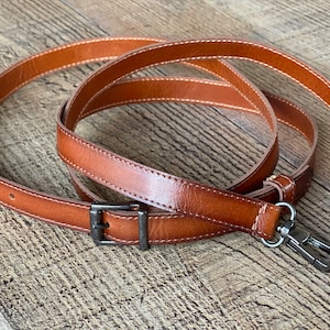 May include: A tan leather bag strap featuring white contrast stitching, an adjustable metal buckle, and two dark gunmetal swivel lobster claw clasps at the ends. The strap is coiled in a circular shape, showcasing the smooth texture of the leather.