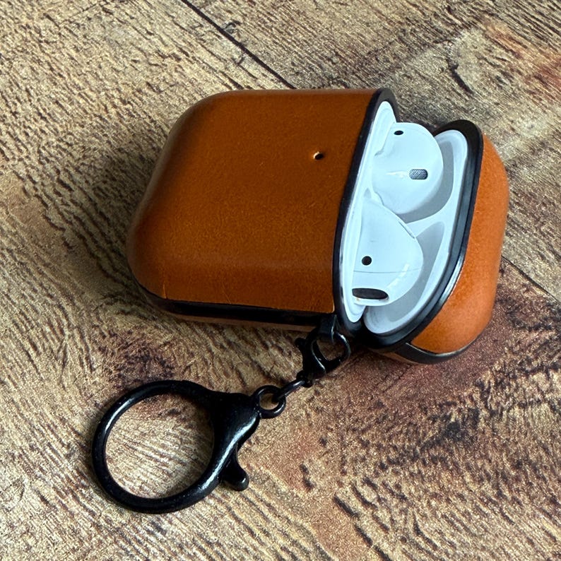 May include: A tan leather protective case for wireless earbuds, featuring a black trim and a black metal lobster clasp keychain attached to the side. The case is open, revealing white wireless earbuds inside the white plastic interior housing.