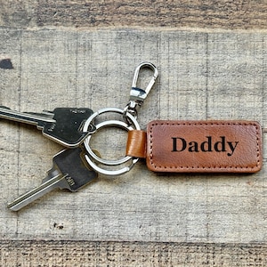 Puede incluir: Un llavero de cuero marrón con la palabra "Daddy" en letras negras. El llavero está unido a un anillo y un cierre de metal plateado, con dos llaves visibles. El fondo es una superficie de madera.