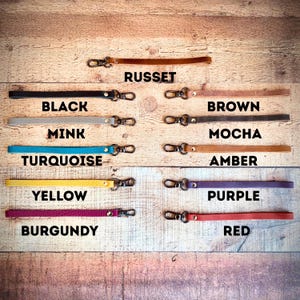 Op de afbeelding: Een verzameling leren polsbandjes in verschillende kleuren, waaronder zwart, bruin en geel. Elke band heeft een metalen sluiting en is gelabeld met de kleurnaam. De bandjes worden gepresenteerd op een houten oppervlak.