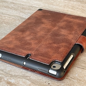 May include: A brown leather tablet case with a textured finish. The case has a magnetic closure and a black frame. The tablet case is designed to protect the device from scratches and damage.