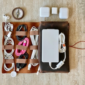May include: A brown leather tech organizer with various white and black charging cables, a white power bank, and a small compass. Also includes white charging blocks and a pair of white earbuds. The organizer is designed to hold and manage electronic accessories.