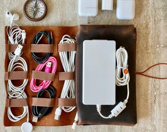 May include: A brown leather tech organizer with various white and black charging cables, a white power bank, and a small compass. Also includes white charging blocks and a pair of white earbuds. The organizer is designed to hold and manage electronic accessories.