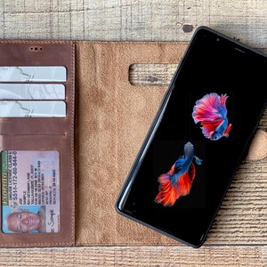 K&ouml;nnte beinhalten: Ein braunes Leder-Brieftaschenetui mit einem Telefon darin. Das Telefon hat einen schwarzen Bildschirm mit einem Bild von zwei roten und blauen Fischen. Die Brieftasche hat drei Steckpl&auml;tze f&uuml;r Karten und ein transparentes Plastikfenster f&uuml;r einen F&uuml;hrerschein. Der Text auf dem F&uuml;hrerschein lautet "Florida Driver License Class E S514-172-80-844-0 Sample 521 GETARNY LANE TALLAHASSEE PL227 Sample REPLACED 08-18".