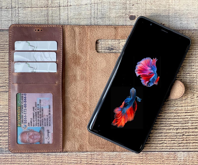 K&ouml;nnte beinhalten: Ein braunes Leder-Brieftaschenetui mit einem Telefon darin. Das Telefon hat einen schwarzen Bildschirm mit einem Bild von zwei roten und blauen Fischen. Die Brieftasche hat drei Steckpl&auml;tze f&uuml;r Karten und ein transparentes Plastikfenster f&uuml;r einen F&uuml;hrerschein. Der Text auf dem F&uuml;hrerschein lautet "Florida Driver License Class E S514-172-80-844-0 Sample 521 GETARNY LANE TALLAHASSEE PL227 Sample REPLACED 08-18".