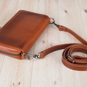May include: A rectangular cognac-colored leather crossbody bag with a smooth finish and a dark metal zipper closure. The bag features a detachable, adjustable matching leather shoulder strap connected by dark metal swivel clasps and rivets.
