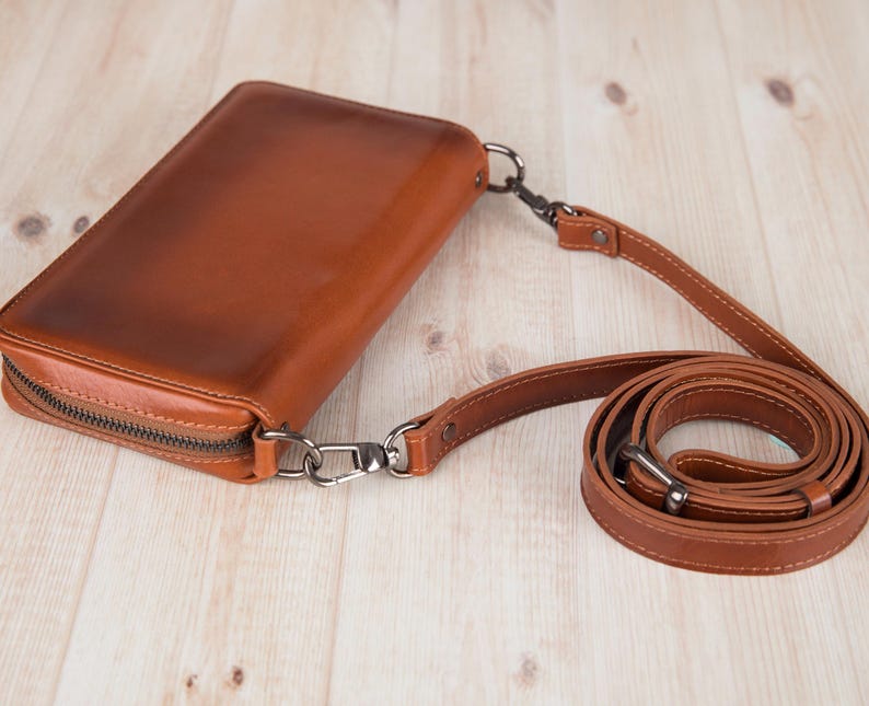 May include: A rectangular cognac-colored leather crossbody bag with a smooth finish and a dark metal zipper closure. The bag features a detachable, adjustable matching leather shoulder strap connected by dark metal swivel clasps and rivets.