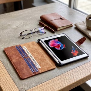 May include: A brown leather tablet case with a tablet and stylus, a brown leather notebook, a pen, and eyeglasses on a table. The tablet displays a colorful fish image. The case has slots for cards and cash.
