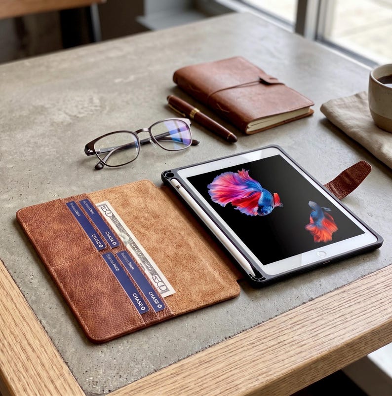 May include: A brown leather tablet case with a tablet and stylus, a brown leather notebook, a pen, and eyeglasses on a table. The tablet displays a colorful fish image. The case has slots for cards and cash.