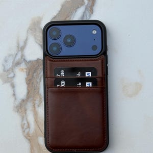 Peut inclure: Une coque de smartphone avec un porte-cartes en cuir marron fonc&eacute; fix&eacute; au dos. La coque poss&egrave;de un cadre noir entourant un module photo bleu avec trois objectifs. Deux cartes bancaires noires sont ins&eacute;r&eacute;es dans les fentes en cuir.