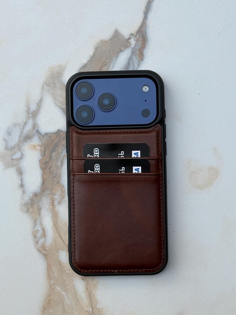 Peut inclure: Une coque de smartphone avec un porte-cartes en cuir marron fonc&eacute; fix&eacute; au dos. La coque poss&egrave;de un cadre noir entourant un module photo bleu avec trois objectifs. Deux cartes bancaires noires sont ins&eacute;r&eacute;es dans les fentes en cuir.