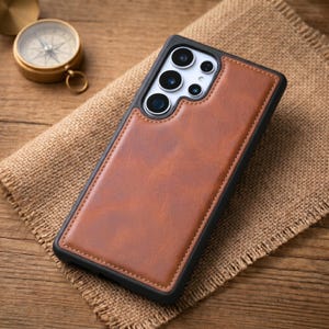 May include: A rectangular smartphone case featuring a smooth brown leather-like back panel with visible stitching along the edges, set within a black frame that includes a cutout for a multi-lens camera array on the upper left side.