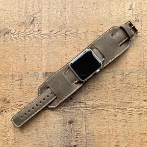 May include: A gray leather watch band with white stitching and a silver watch face. The band has a wide cuff design and a buckle closure. The watch band is shown on a wooden surface.