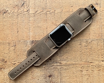 May include: A gray leather watch band with white stitching and a silver watch face. The band has a wide cuff design and a buckle closure. The watch band is shown on a wooden surface.