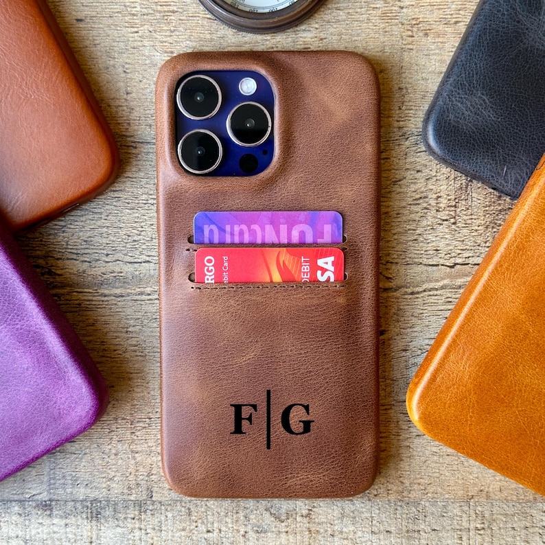 Peut inclure: Une coque de t&eacute;l&eacute;phone en cuir marron avec une fente pour cartes contenant deux cartes de cr&eacute;dit. La coque pr&eacute;sente une d&eacute;coupe pour l'appareil photo et les initiales F G imprim&eacute;es en noir s&eacute;par&eacute;es par une ligne verticale.